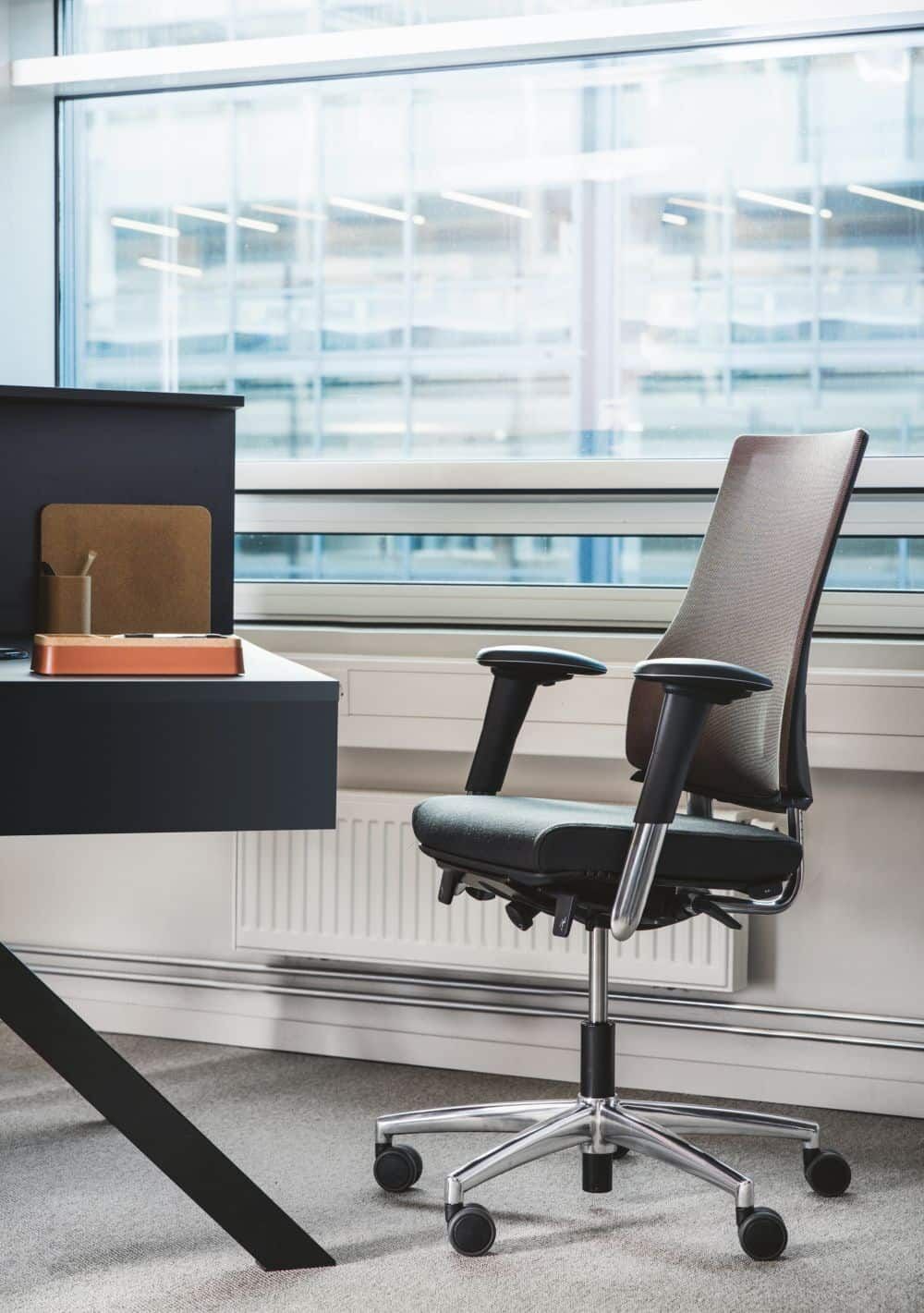 Ergonomischer Bürostuhl mit Netzrückenlehne und Rollen neben einem modernen Schreibtisch vor einem großen Fenster.