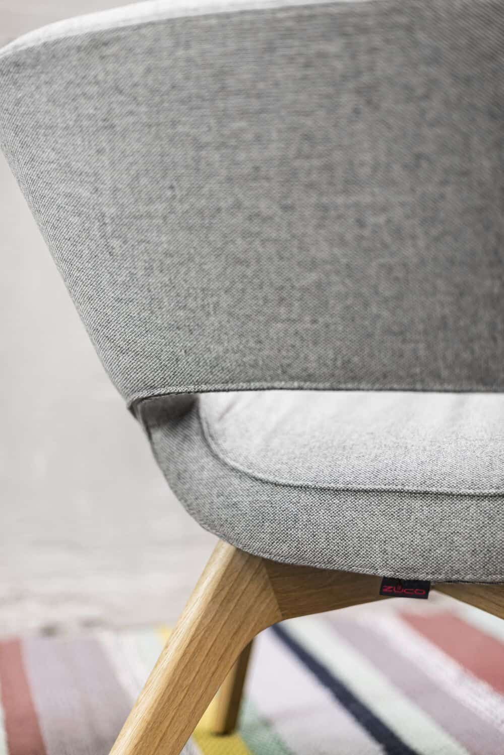 Nahaufnahme eines ergonomischen Stuhls mit grauem Stoffbezug und Holzbeinen auf einem bunten Teppich.