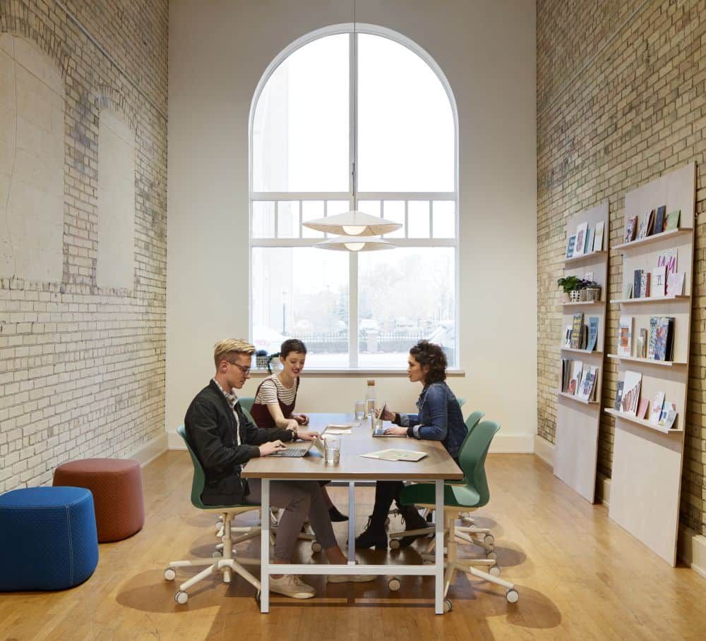 Drei Personen arbeiten an einem Tisch in einem modernen, hellen Büro mit großen Fenstern und Bücherregalen.
