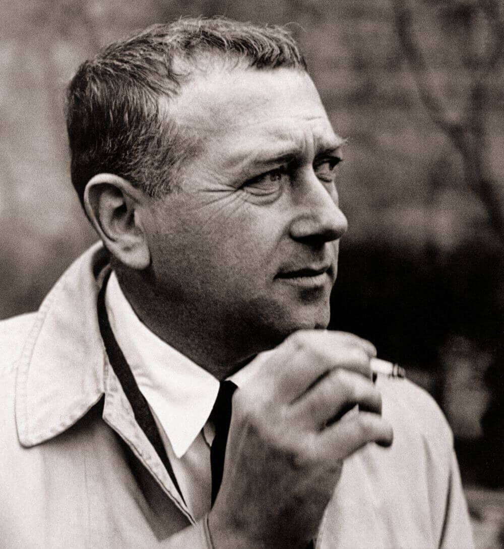 Schwarzweißfoto eines Mannes in einem Trenchcoat, der seitlich schaut und eine Zigarette hält.