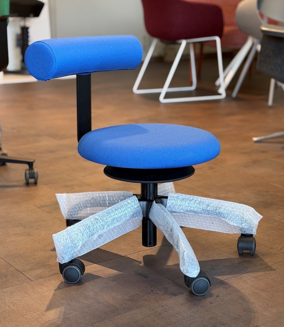 Blauer ergonomischer Bürostuhl mit Polsterung und Rollen, teils in Schutzfolie, auf Holzboden ausgestellt.