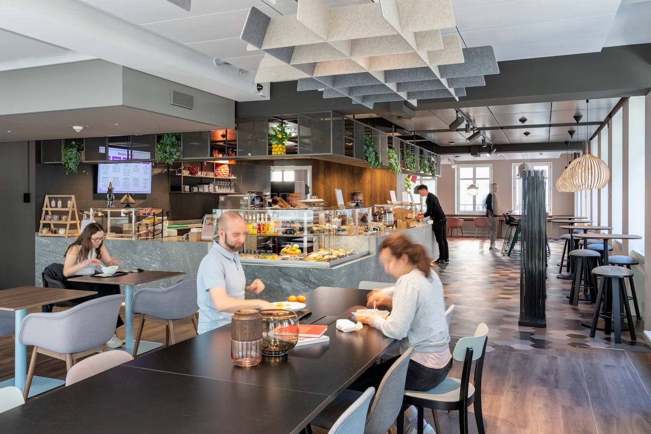 Modernes Café mit Holz- und Stilelementen; Menschen sitzen und genießen Mahlzeiten, während Barista arbeitet.