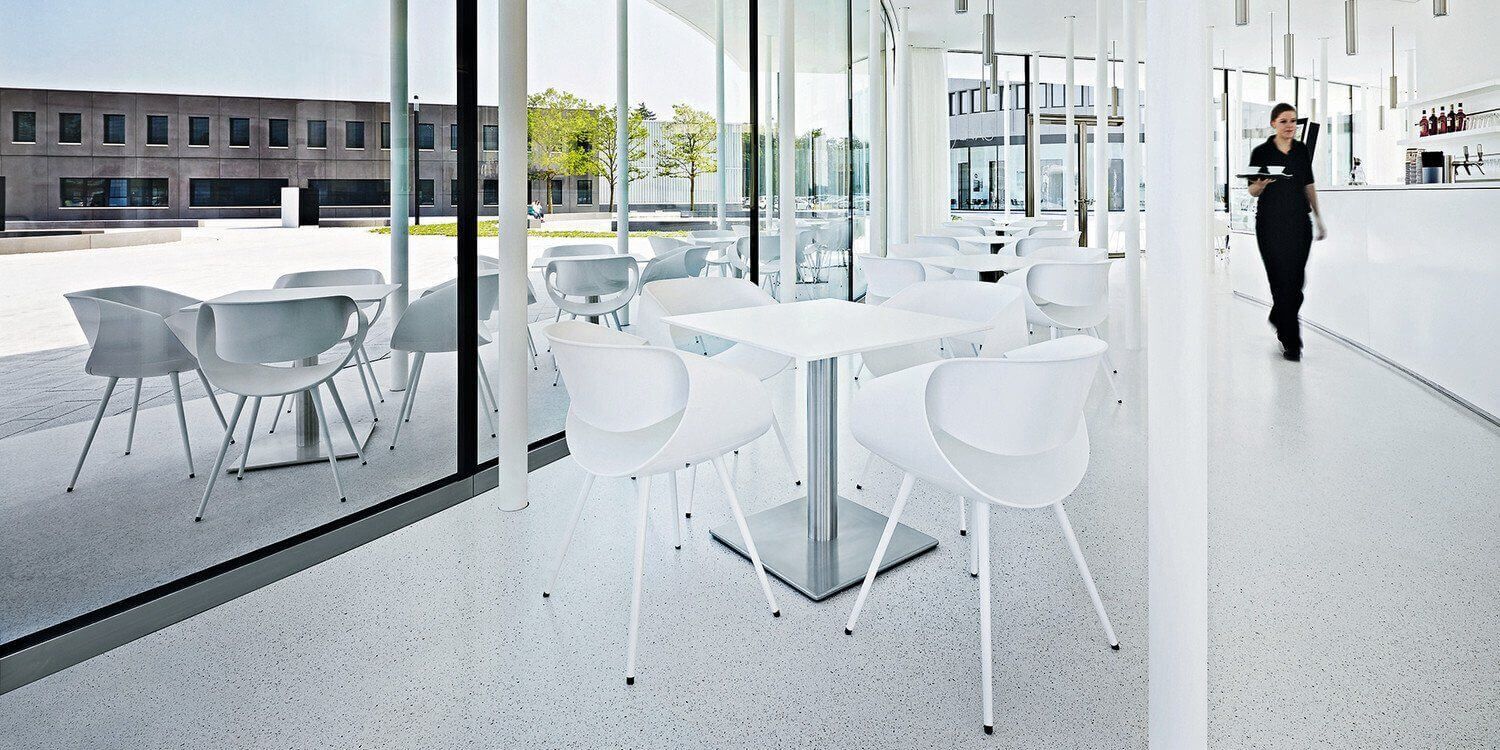 Moderner, heller Cafébereich mit weißen ergonomischen Stühlen und Tischen, Bedienung im Hintergrund.