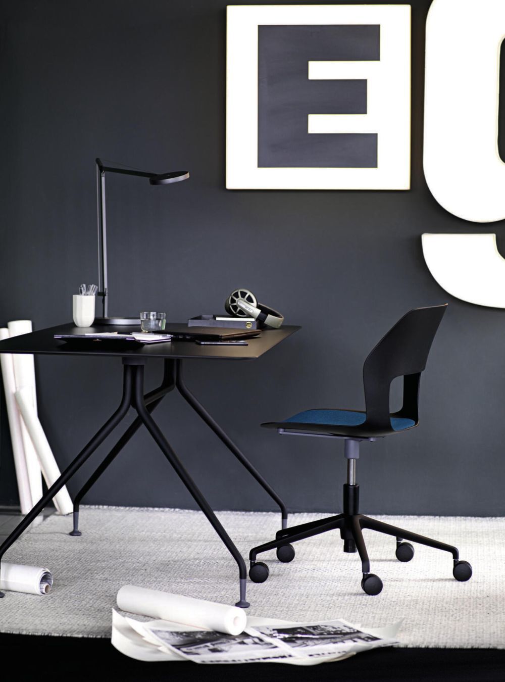 Moderner Schreibtisch mit Lampe und ergonomischem Bürostuhl vor dunkler Wand mit großen Buchstaben.