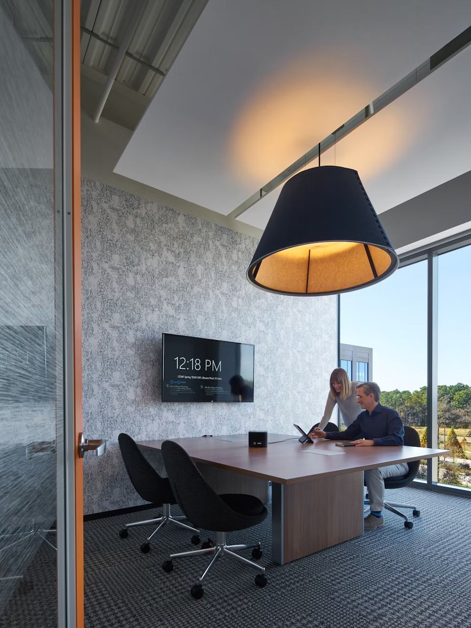 Besprechungsraum mit großem Holztisch, ergonomischen Stühlen, modernem Bildschirm und zwei Personen in Besprechung.