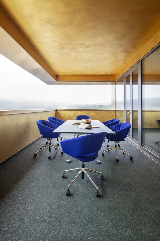 Moderner Balkon mit blau gepolsterten Bürostühlen und Tisch, umgeben von Glaswänden, mit Aussicht auf die Landschaft.