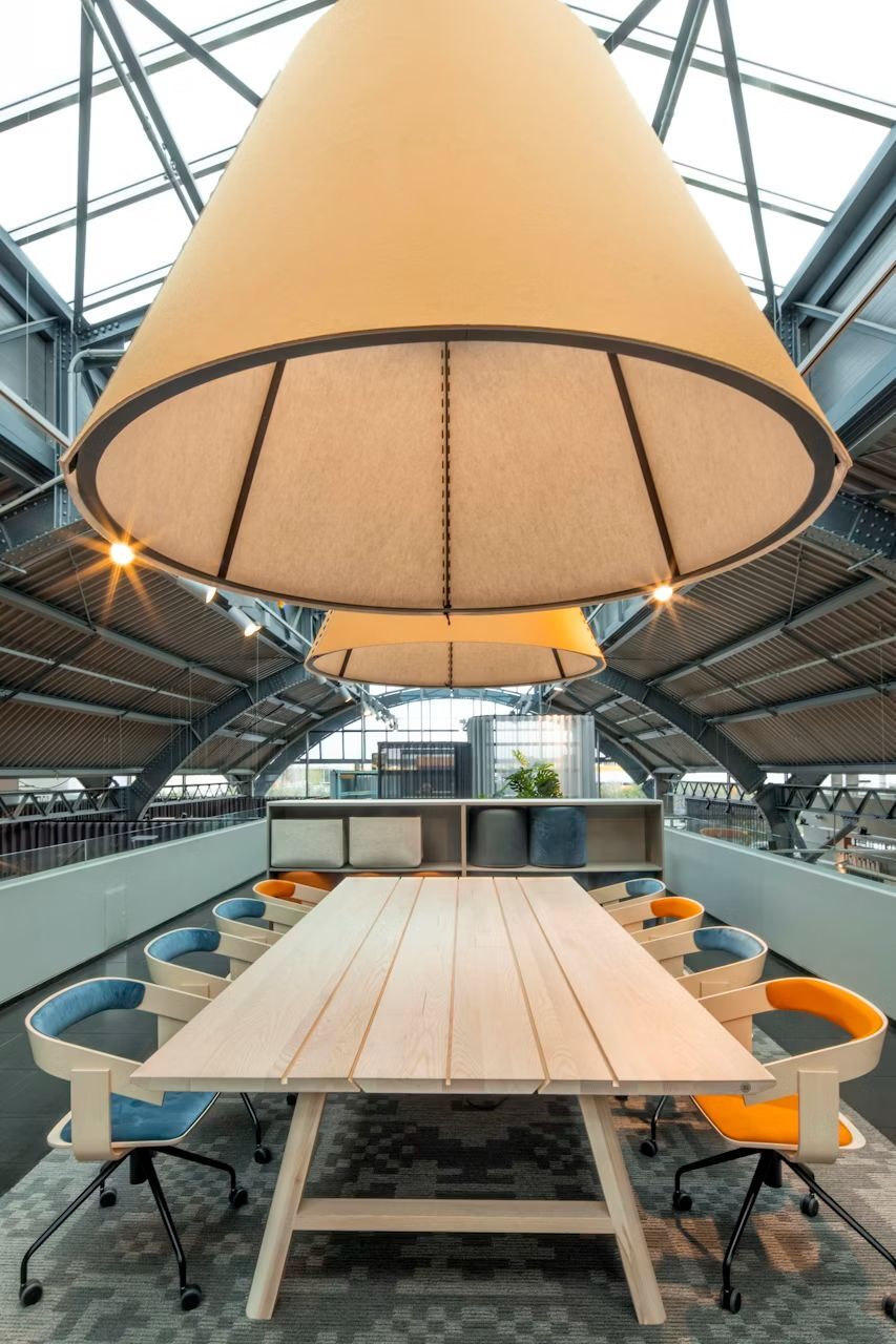 Moderner Konferenztisch mit bunten ergonomischen Stühlen, darunter große Lampenschirme in einem industriellen Raum.