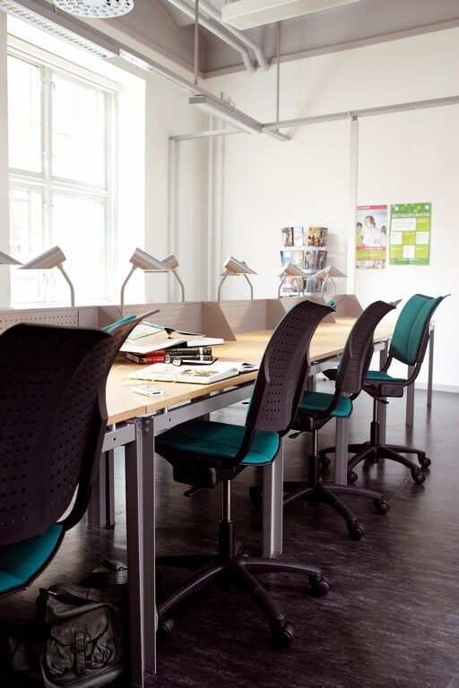 Ergonomische Bürostühle an einem modernen Arbeitstisch in einem sonnigen Büro mit Laptops und Leuchten.