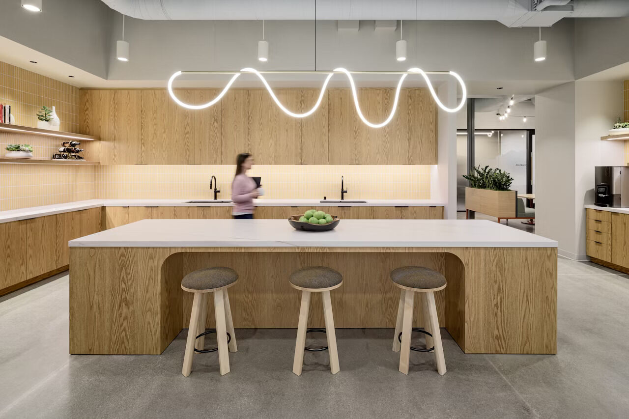 Moderne Küche mit Holzvertäfelung, zentralem Tisch und wellenförmiger Deckenleuchte. Person im Hintergrund.