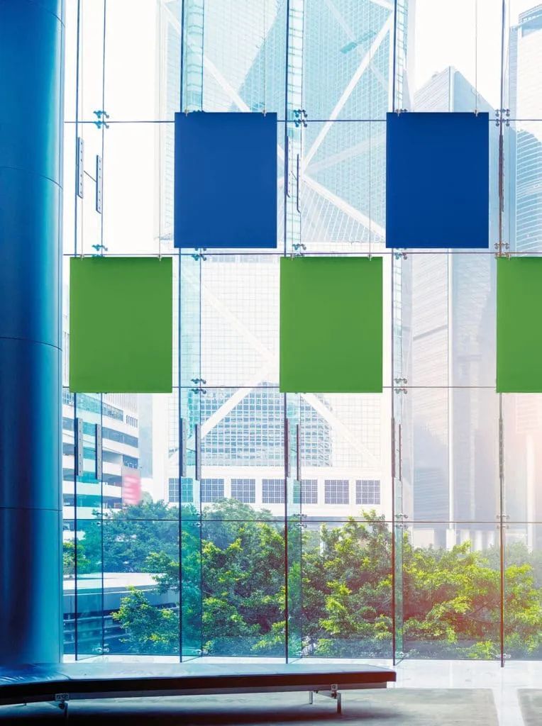 Blick durch große Glasfenster mit blauen und grünen Paneelen auf städtische Architektur und Bäume draußen.