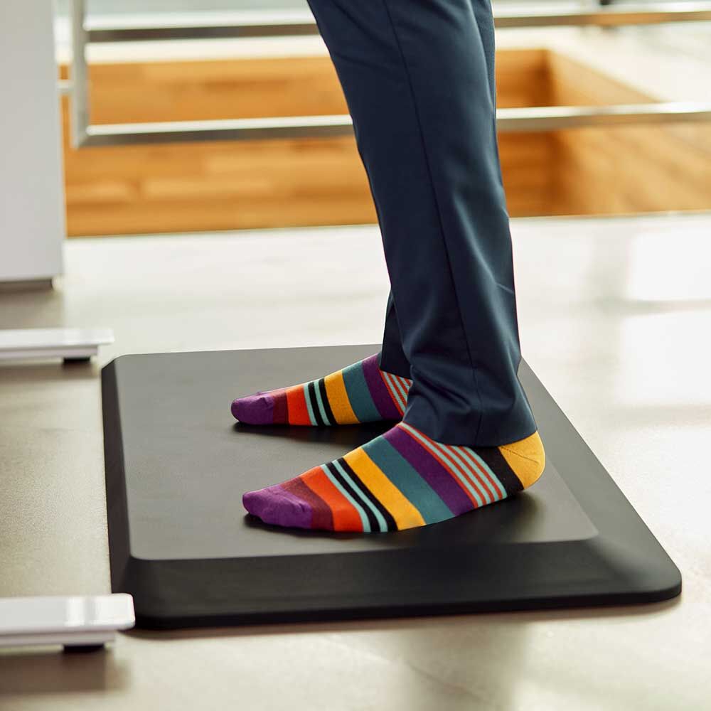 Person steht auf ergonomischer Anti-Ermüdungsmatte, trägt bunte Socken und blaue Hose.