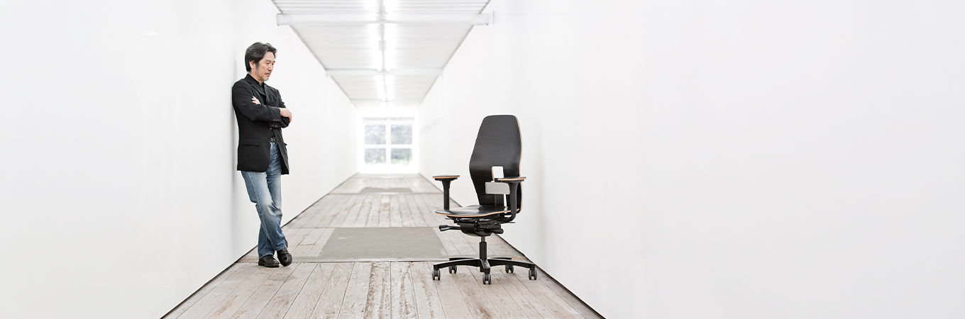 Mann steht hinter einem ergonomischen Bürostuhl an einem Holztisch vor einer Backsteinmauer.
