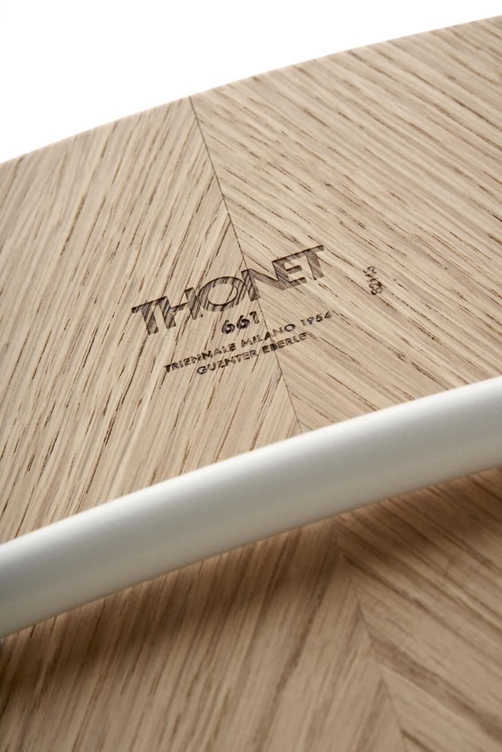 Nahaufnahme eines Holzschriftzugs „THONET“ auf einer hellen Holzoberfläche, teilweise von einem weißen Rohr verdeckt.