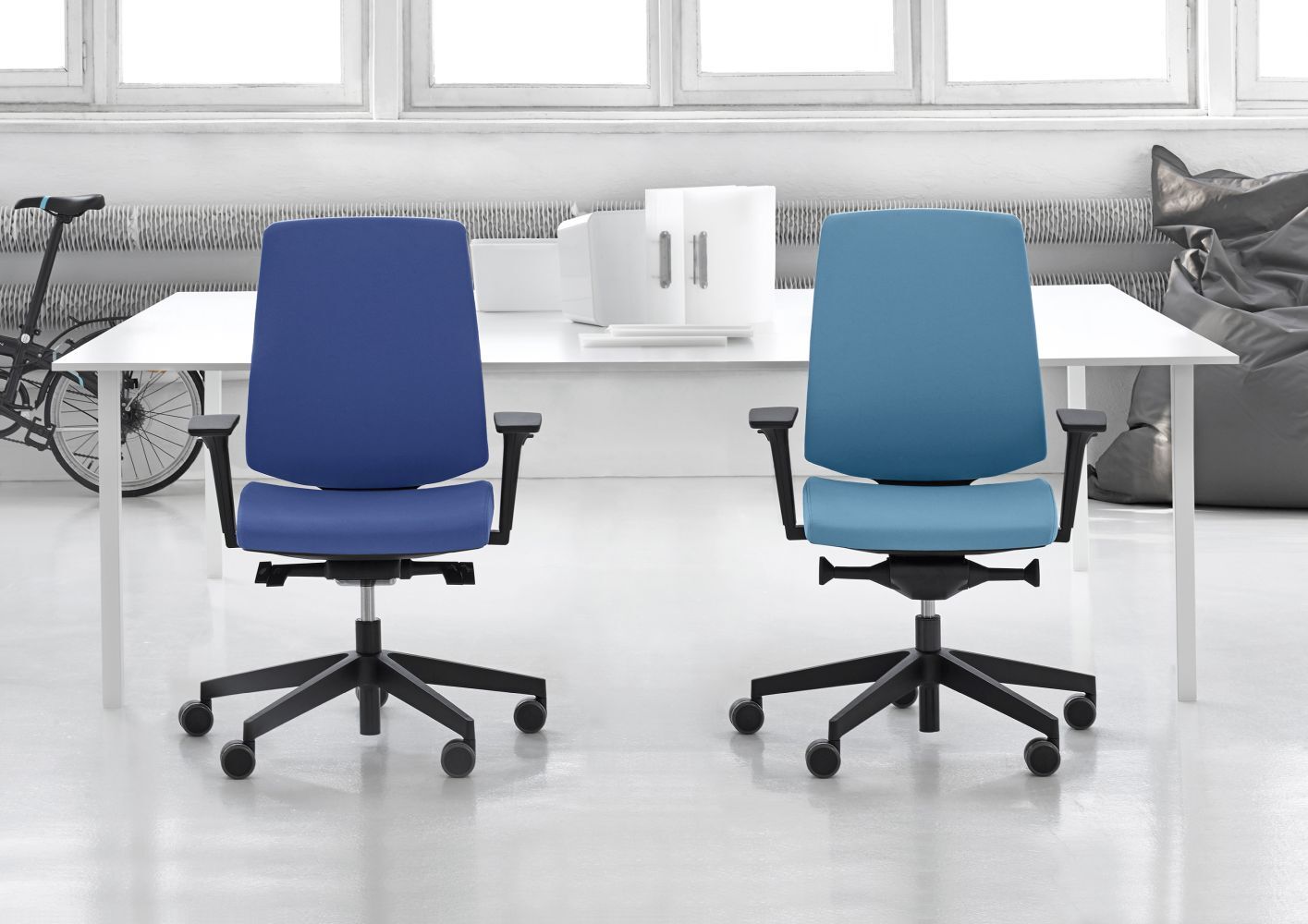Zwei ergonomische Bürostühle in blau vor einem weißen Schreibtisch, mit hellem, modernem Hintergrund.