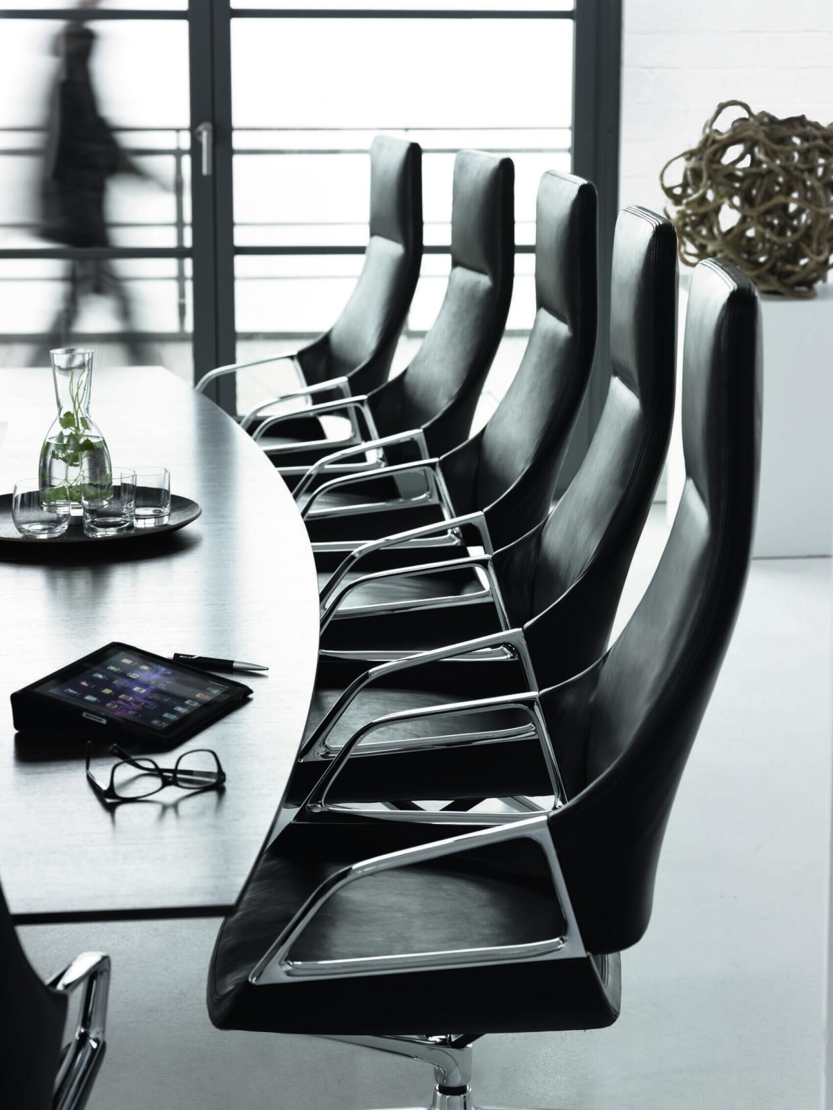Mehrere ergonomische Bürostühle aus Leder an einem Konferenztisch mit Tablett, Brille und Wasserkrug.