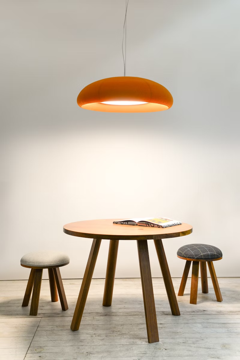 Runder Holztisch mit zwei Hockern, darüber eine orange Hängelampe; auf dem Tisch liegt ein aufgeschlagenes Magazin.