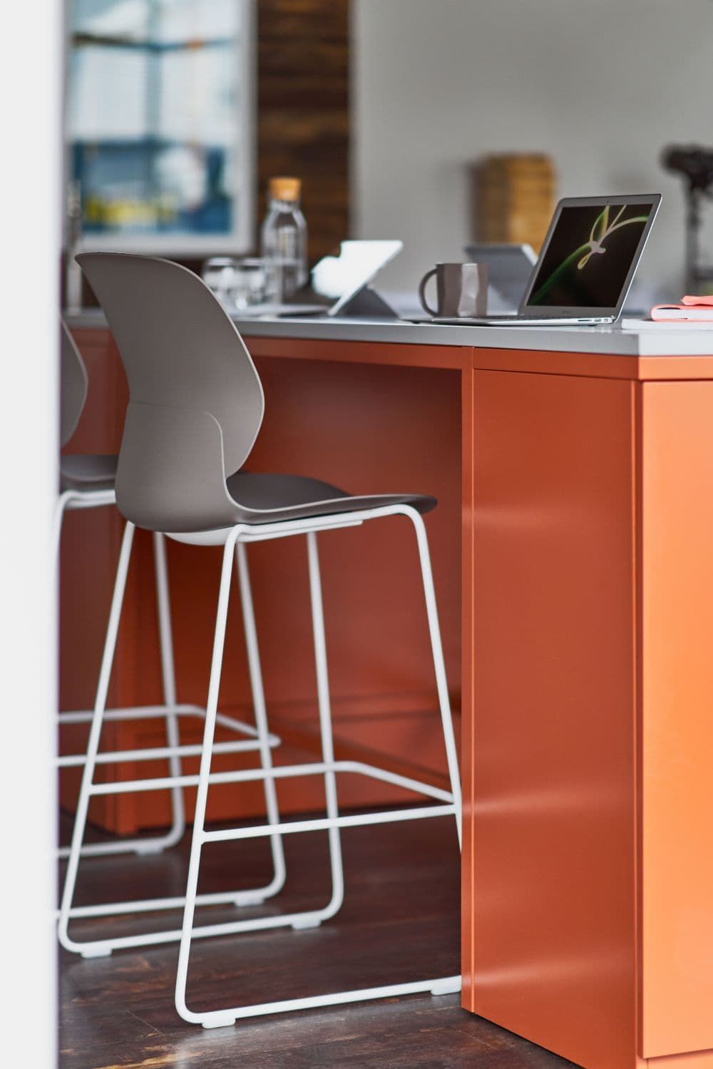 Moderne Kücheninsel mit Laptop, grauen Barhockern und orangefarbenen Oberflächen, ideal für dynamisches Arbeiten.