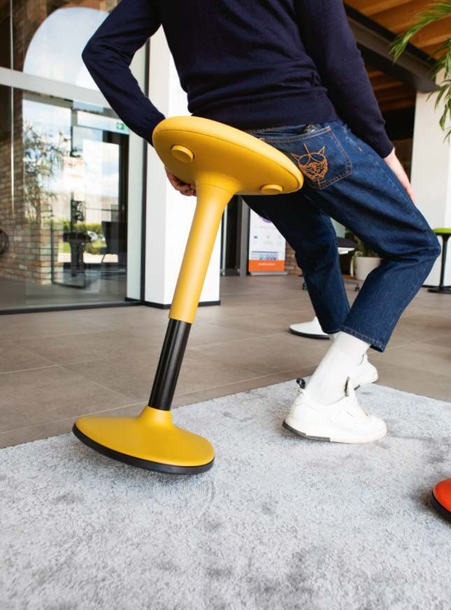 Person sitzt dynamisch auf einem gelben, ergonomischen Hocker auf einem grauen Teppich.