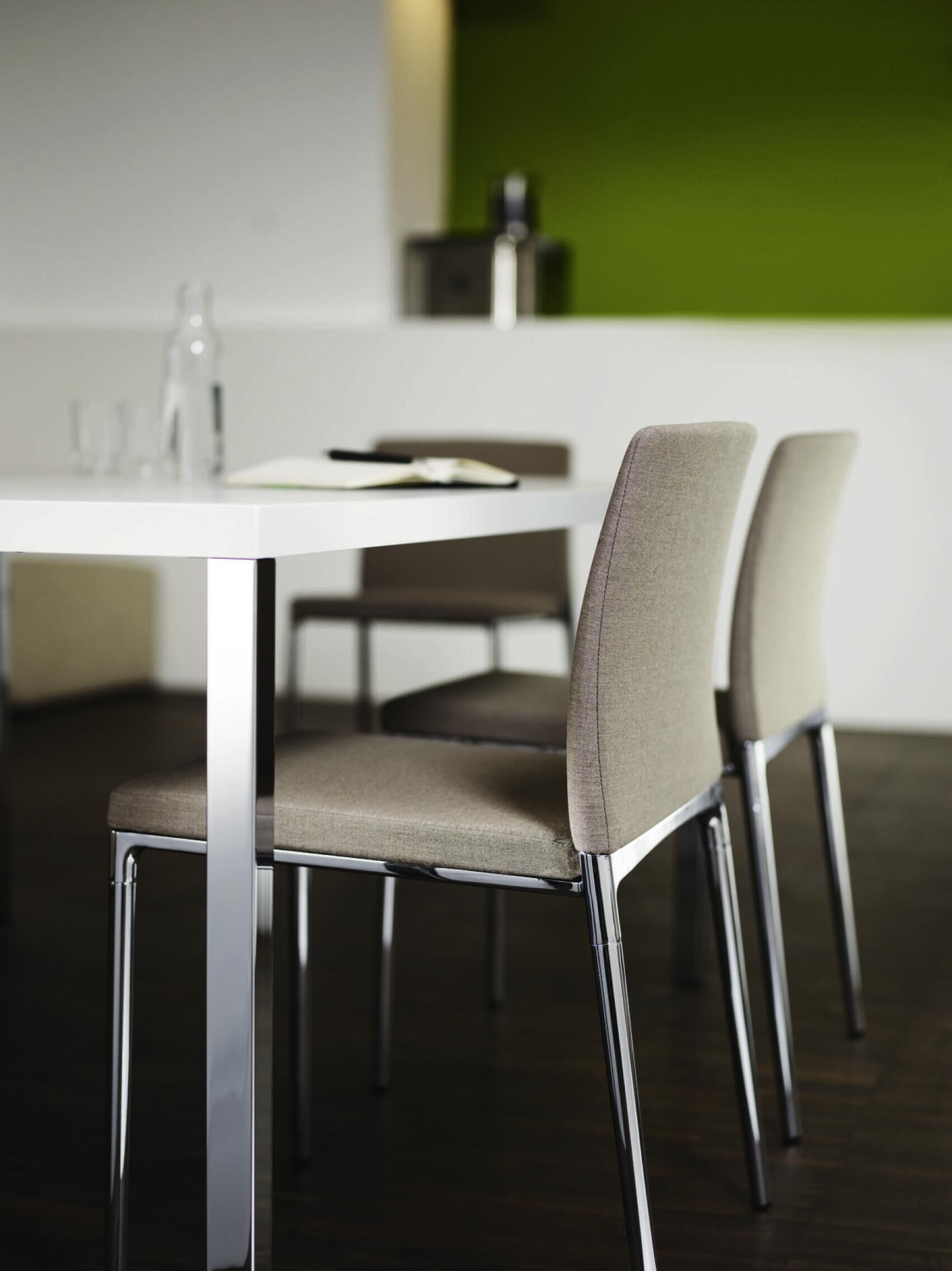 Moderne Stühle mit Metallbeinen an einem hellen Tisch, vor einer grünen Wand in einem Bürobereich.