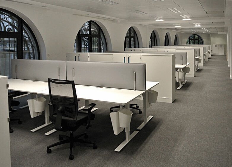 Moderne, offene Bürofläche mit ergonomischen Drehstühlen und weißen Tischen vor großen Bogenfenstern.