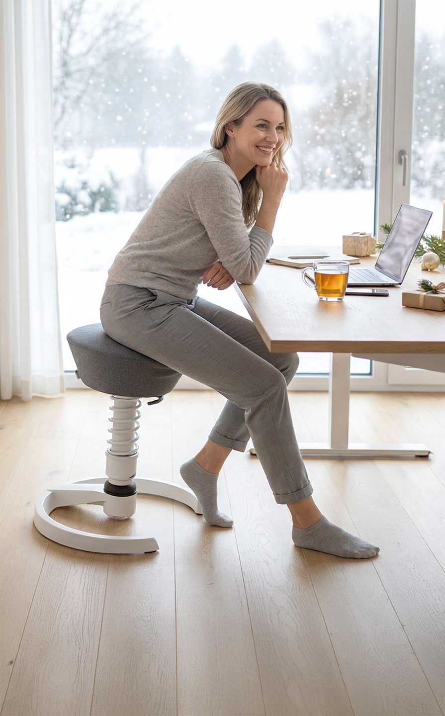 Frau sitzt auf ergonomischem Hocker an Schreibtisch mit Laptop, umgeben von weihnachtlicher Dekoration.