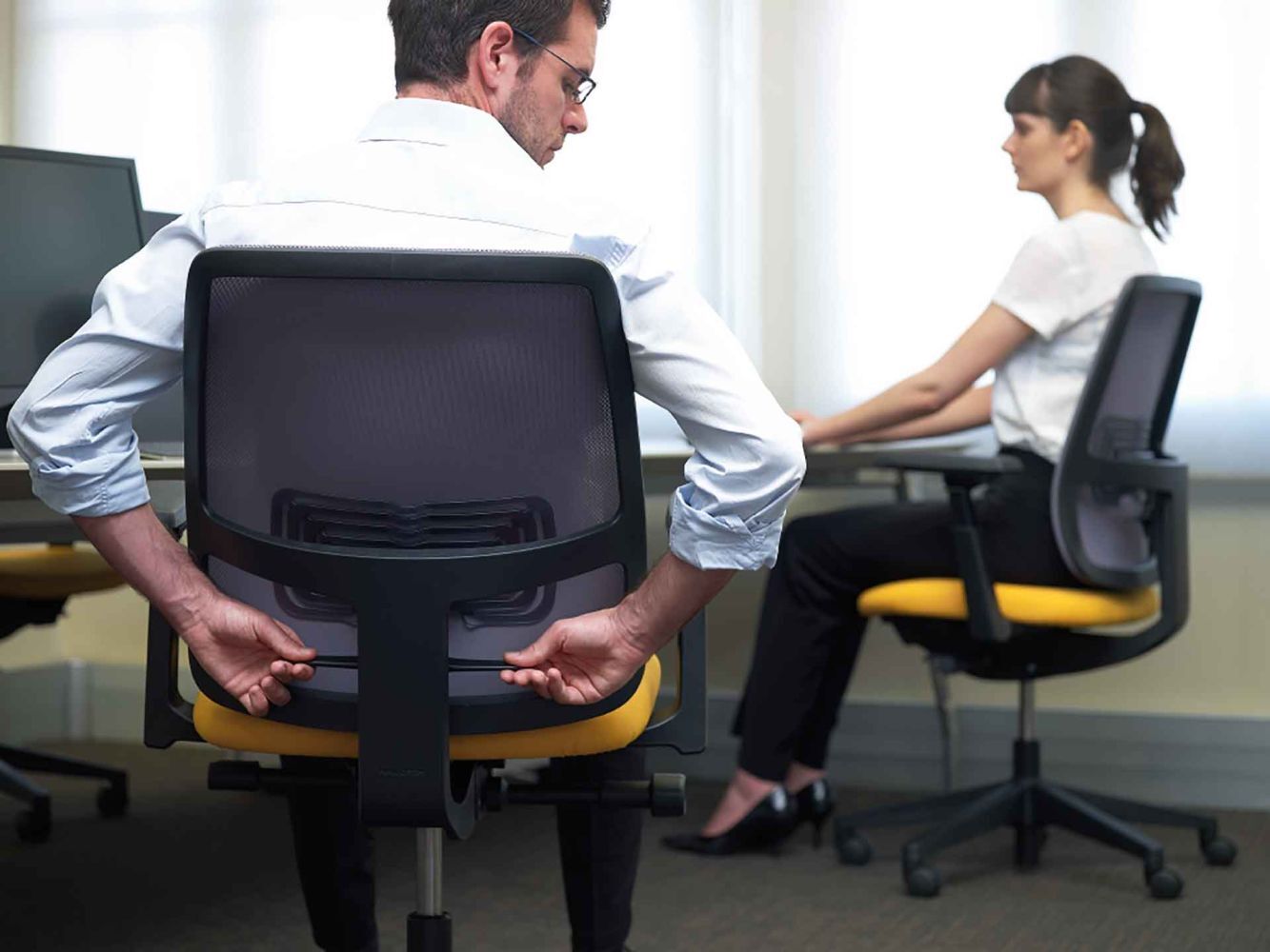 Ein Mann justiert einen ergonomischen Bürostuhl, während eine Frau an einem Schreibtisch sitzt.