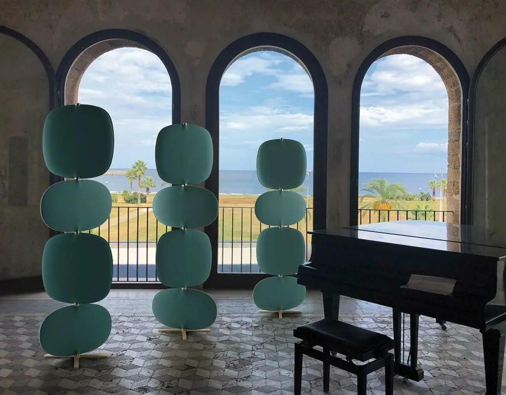 Raum mit Klavier, drei grünen Raumteilern und Blick auf den Strand durch große Fensterbögen.