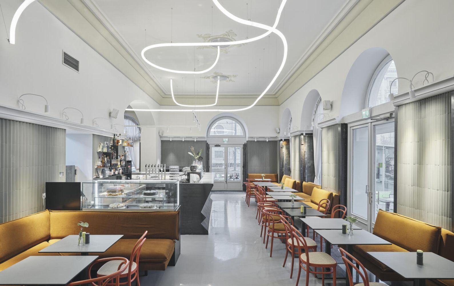 Modernes Café mit eleganter Beleuchtung, gelben Sitzbänken, roten Stühlen und einer Theke mit Gebäck.