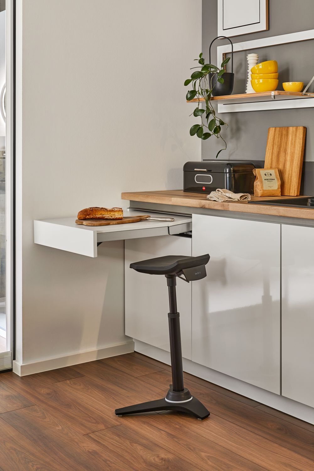 Ergonomischer Hocker in moderner Küche neben ausziehbarem Tisch mit Brot, Pflanzen und Küchenutensilien.