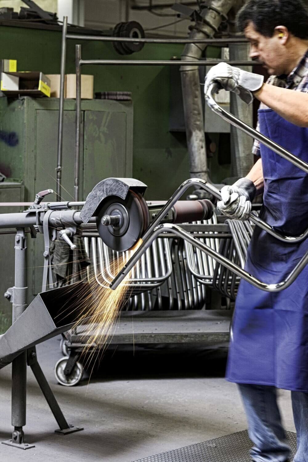 Arbeiter schleift Metallstuhlrahmen in einer Werkstatt mit Funken sprühender Schleifmaschine, zeigt Handwerkskunst.