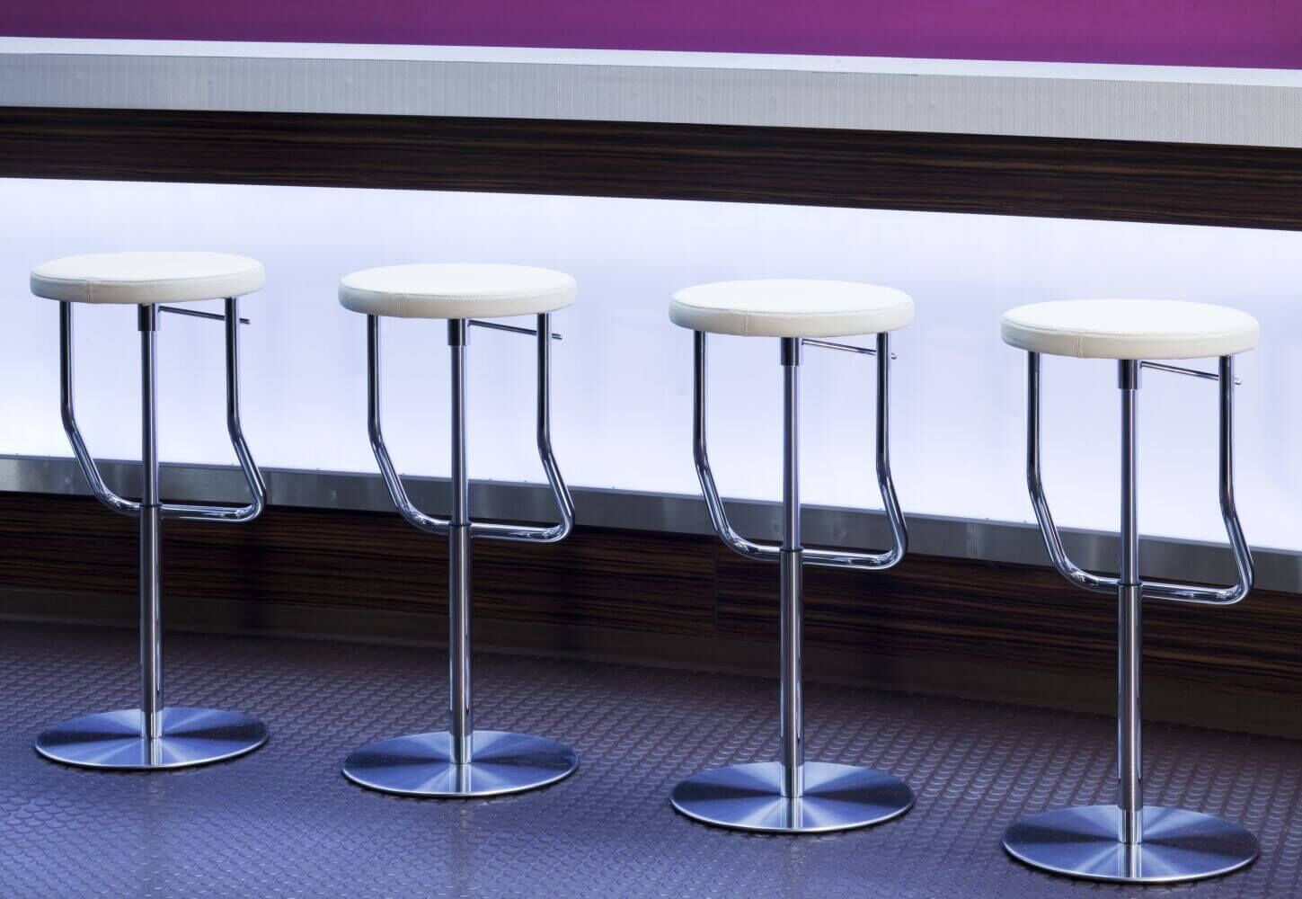 Vier ergonomische Barhocker mit chromfarbenen Gestellen und weißen Sitzflächen vor einer modernen Theke.