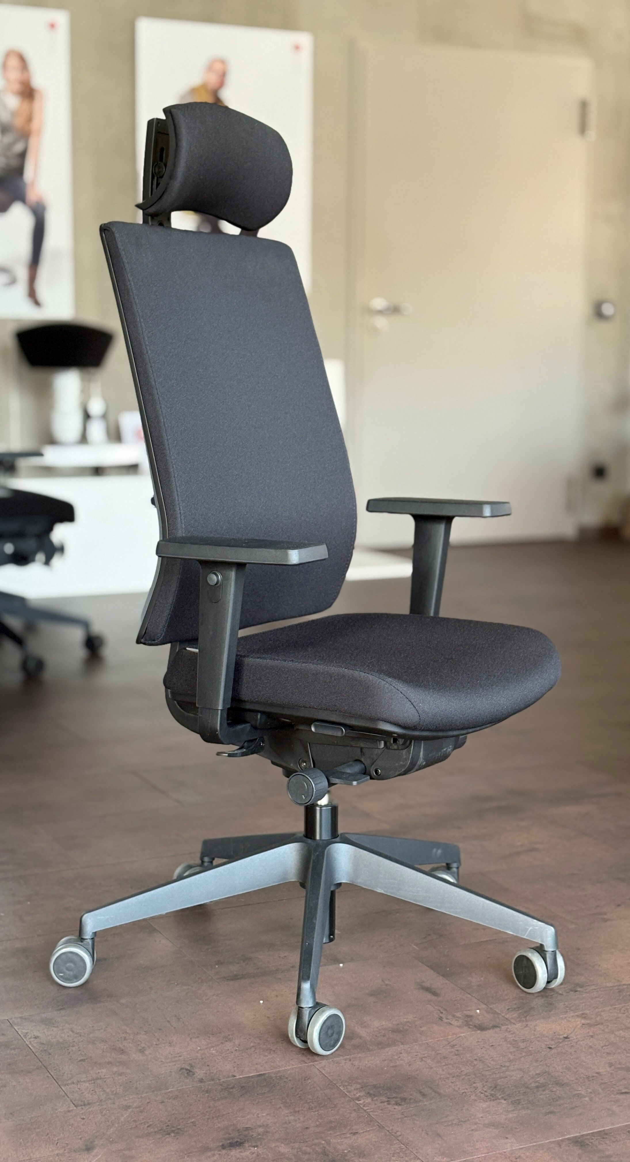 Ergonomischer schwarzer Bürostuhl mit verstellbarer Kopfstütze, Armlehnen und Rollen auf Holzfußboden.
