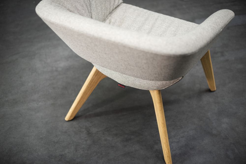 Grauer ergonomischer Stuhl mit gepolstertem Sitz und Holzbeinen auf dunklem Boden, schräg von oben fotografiert.