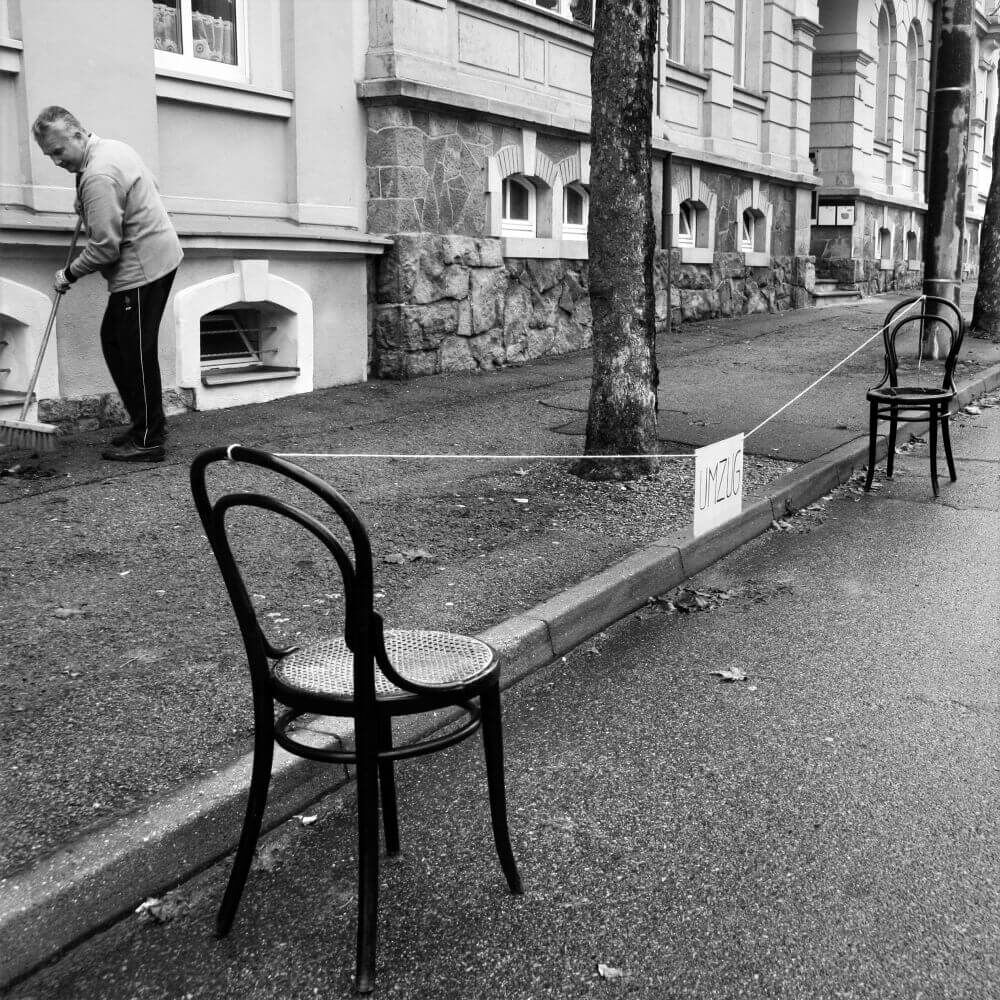 Älterer Mann fegt Gehweg; Stühle sperren Bereich ab mit Schild 'Umzug' auf Bürgersteig.