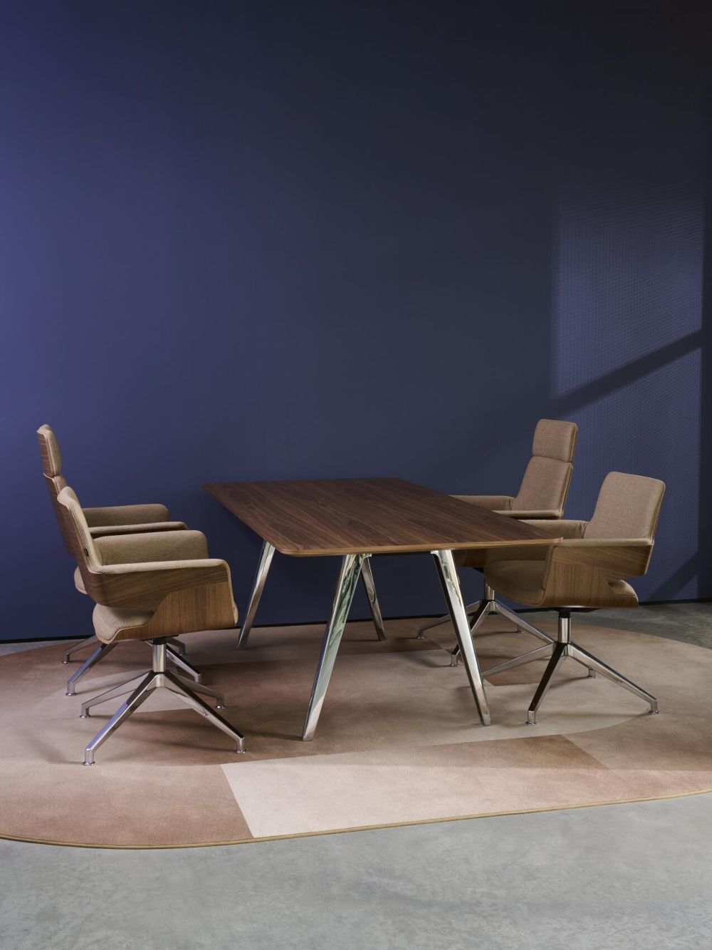 Moderner Konferenztisch mit vier ergonomischen Drehstühlen auf einem gemusterten Teppich, vor einer blauen Wand.