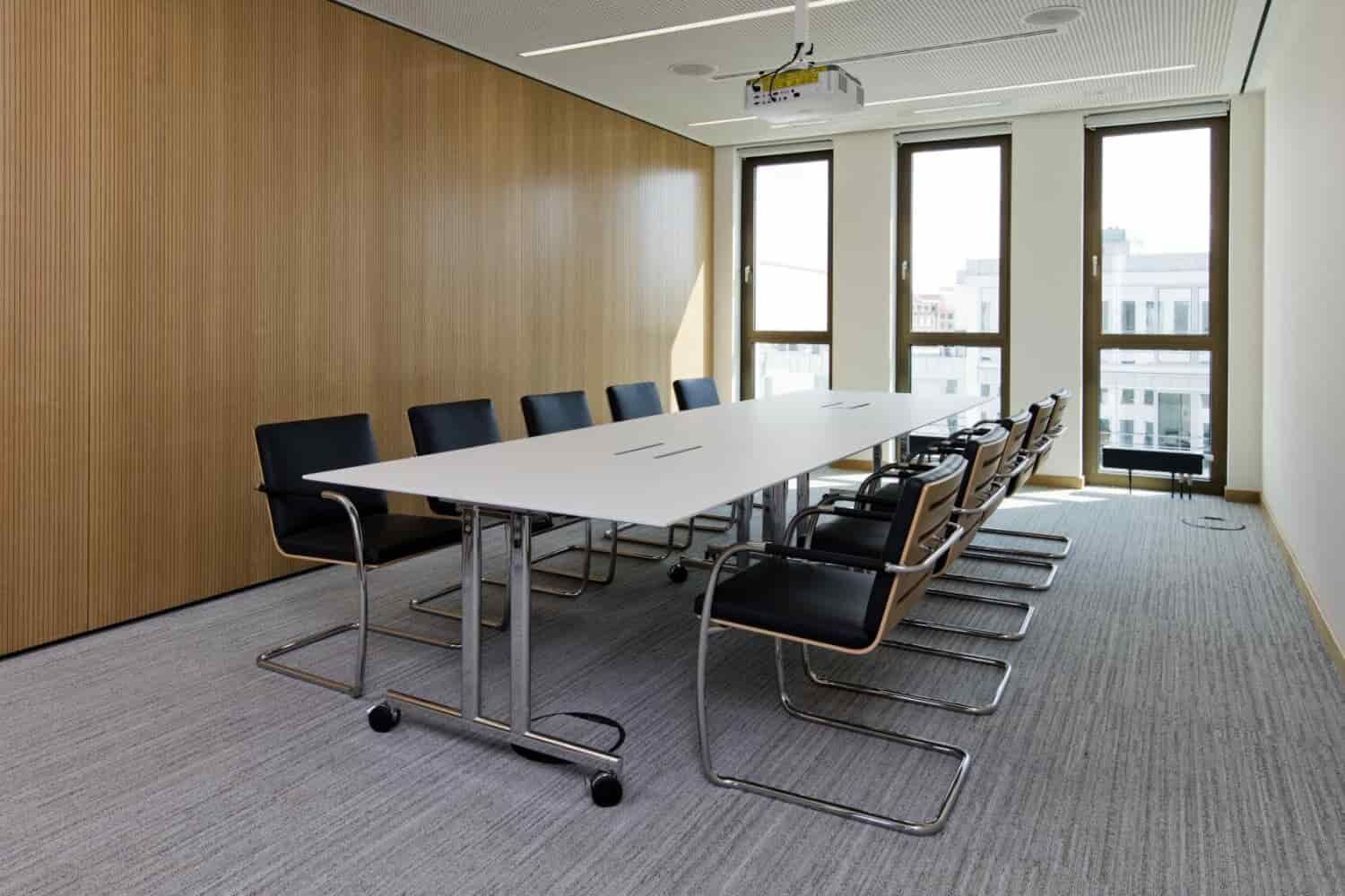Moderner Konferenzraum mit langem weißen Tisch und schwarzen ergonomischen Stühlen vor großen Fenstern.