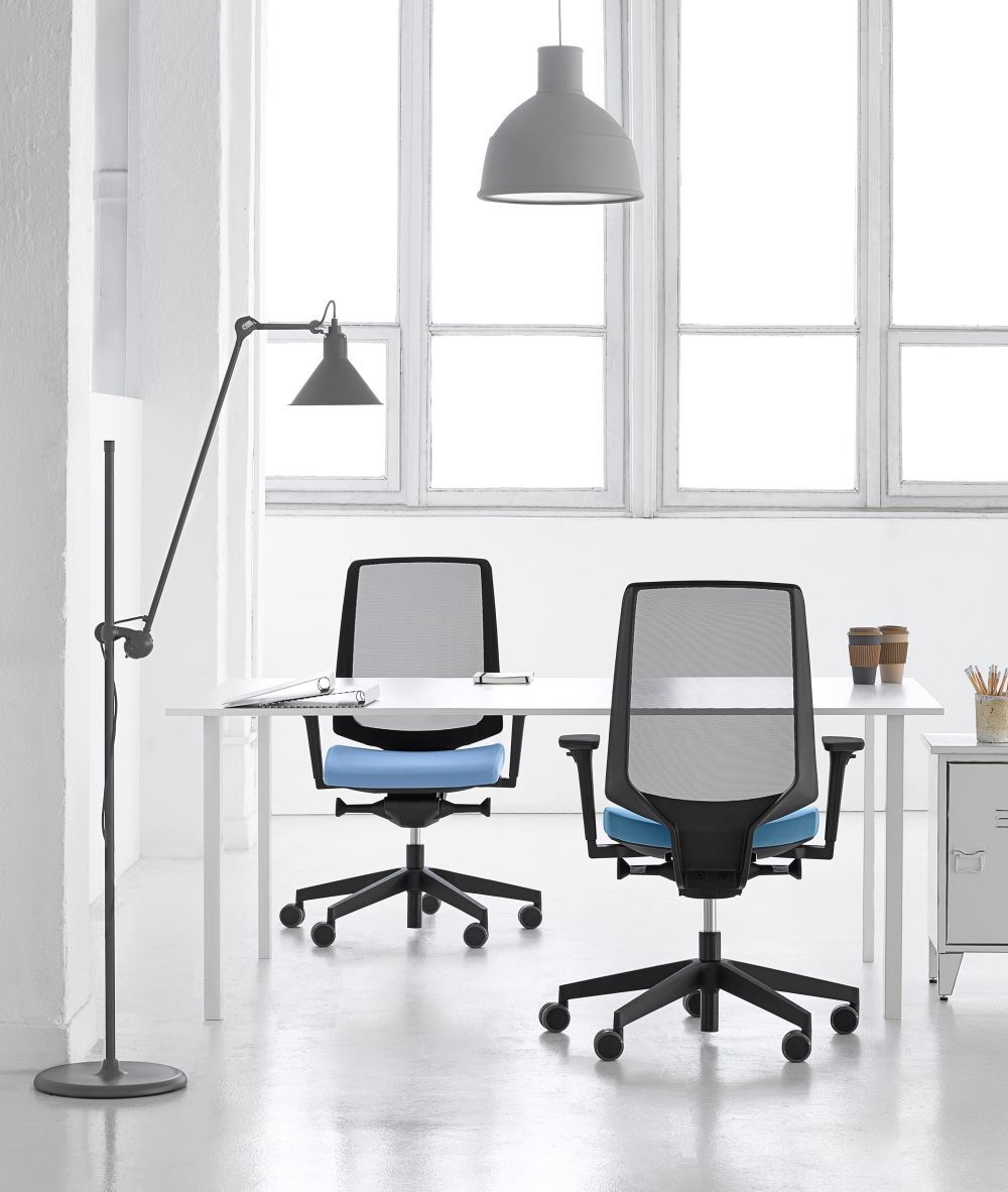 Zwei ergonomische Bürostühle an einem weißen Tisch in einem modernen, hellen Büro mit großen Fenstern.