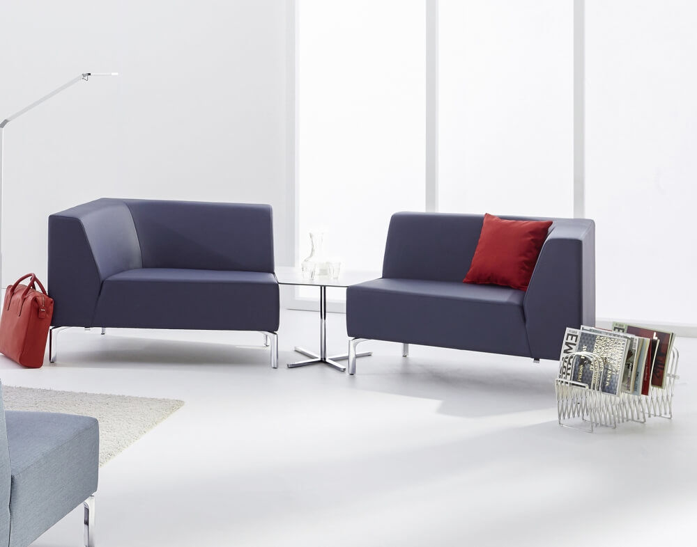 Moderner Loungebereich mit zwei dunkelgrauen Sofas, einem Glastisch, rotem Kissen und Zeitschriftenständer.