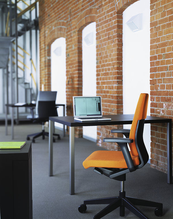Ergonomischer Bürostuhl in Orange vor einem Schreibtisch mit Laptop, in modernem Büro mit Backsteinwand.