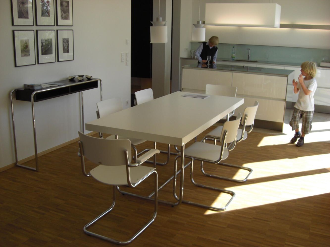 Moderner Essbereich mit weißen Stühlen und Tisch, hell beleuchtet durch Sonnenlicht, Küche im Hintergrund.