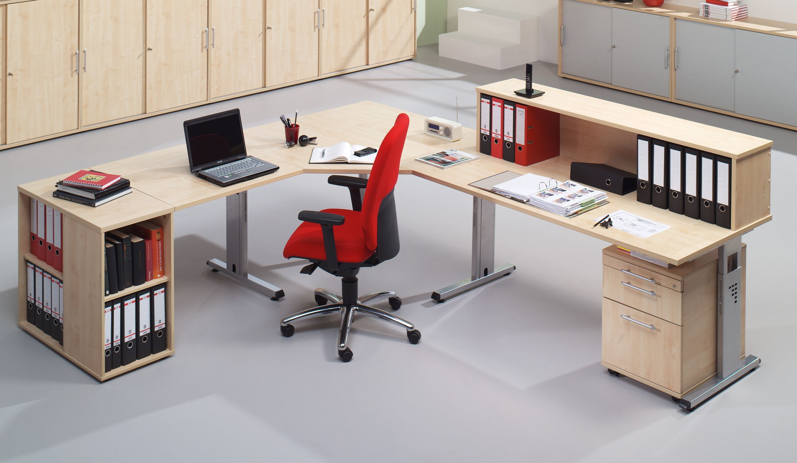 Moderner, ergonomischer Bürostuhl vor einem L-förmigen Schreibtisch mit Laptop, Ordnern und Büroartikeln.