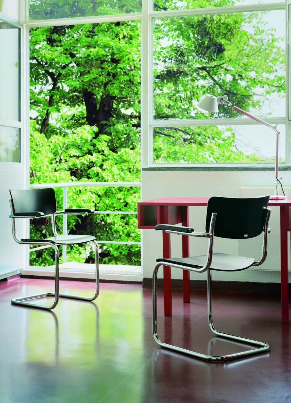 Zwei ergonomische Freischwingerstühle und ein Schreibtisch vor einem großen Fenster mit Blick auf grüne Bäume.