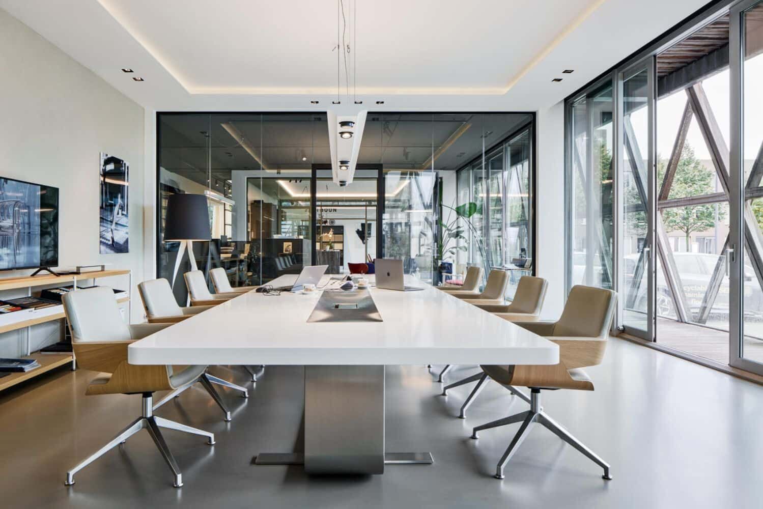 Moderner Konferenzraum mit ergonomischen Drehstühlen um einen langen weißen Tisch, umgeben von Glaswänden.